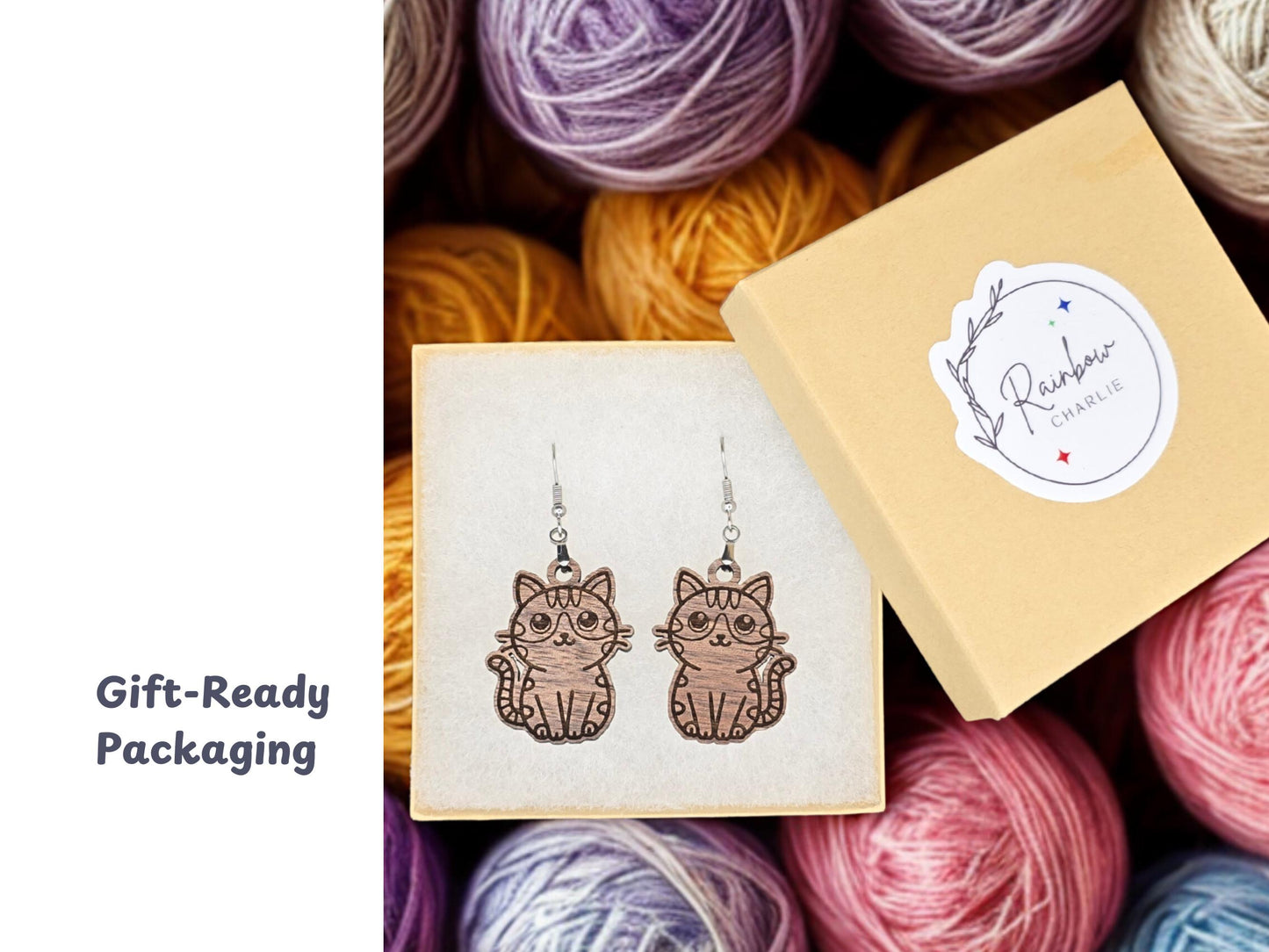 Pair of walnut wood cat earrings inside a cream-colored gift box with a Rainbow Charlie branded sticker on the lid. Colorful yarn balls fill the background. A label stating "Gift-Ready Packaging."