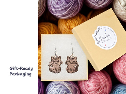 Pair of walnut wood cat earrings inside a cream-colored gift box with a Rainbow Charlie branded sticker on the lid. Colorful yarn balls fill the background. A label stating "Gift-Ready Packaging."