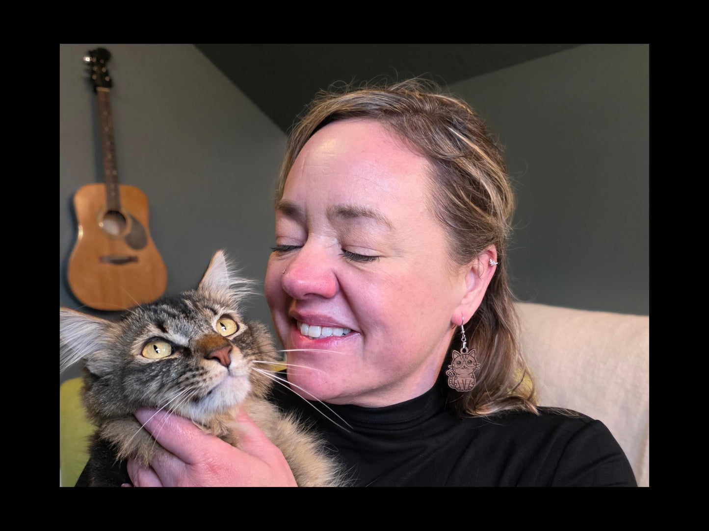 Woman wearing walnut wood cat earrings while holding a fluffy tabby cat. The earring hangs from a nickel-free hook and features an engraved cat design. A guitar hangs on the wall in the background.