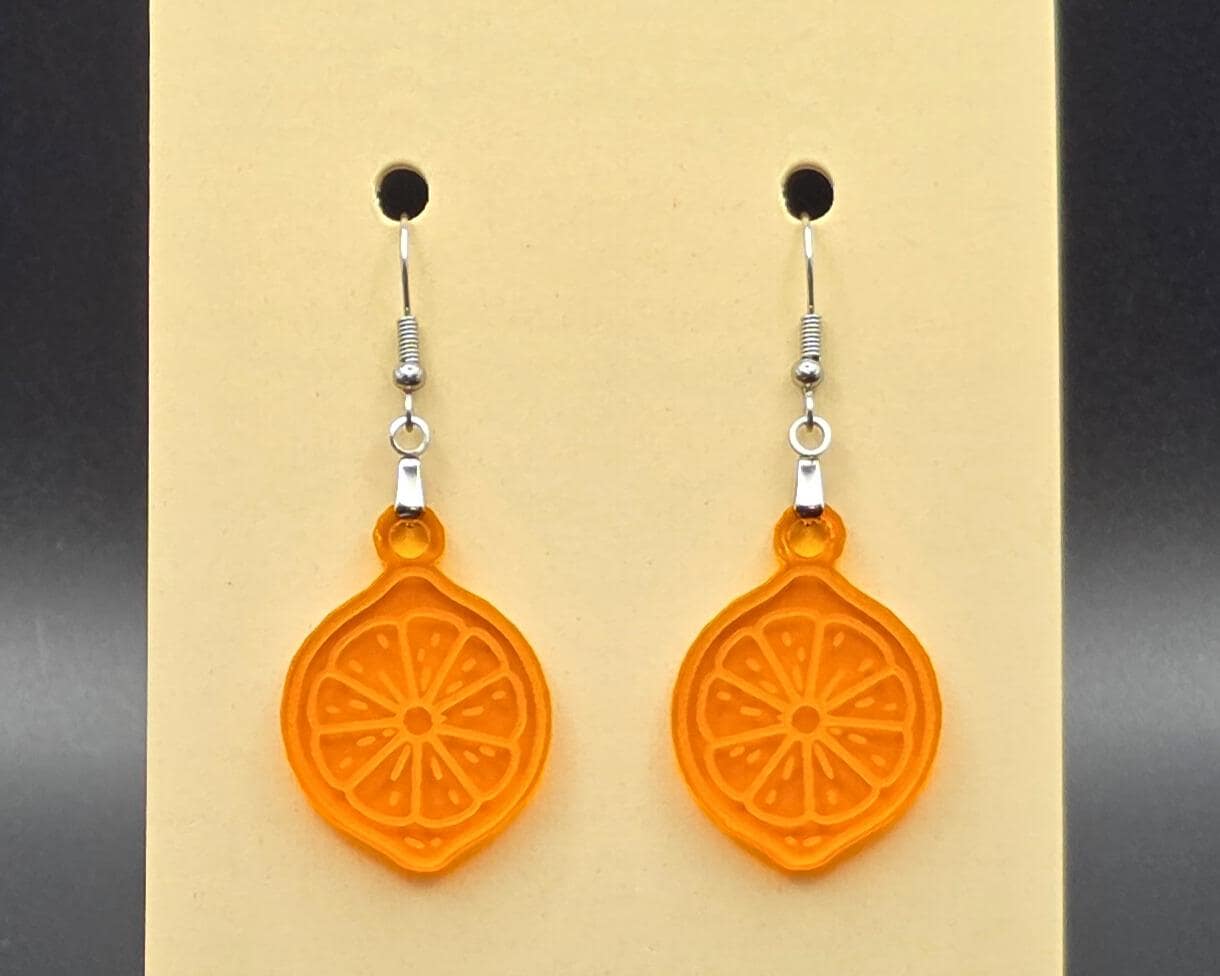 A pair of orange acrylic citrus slice earrings on a beige earring display card. The translucent orange charms resemble detailed orange slices with engraved wedge patterns.