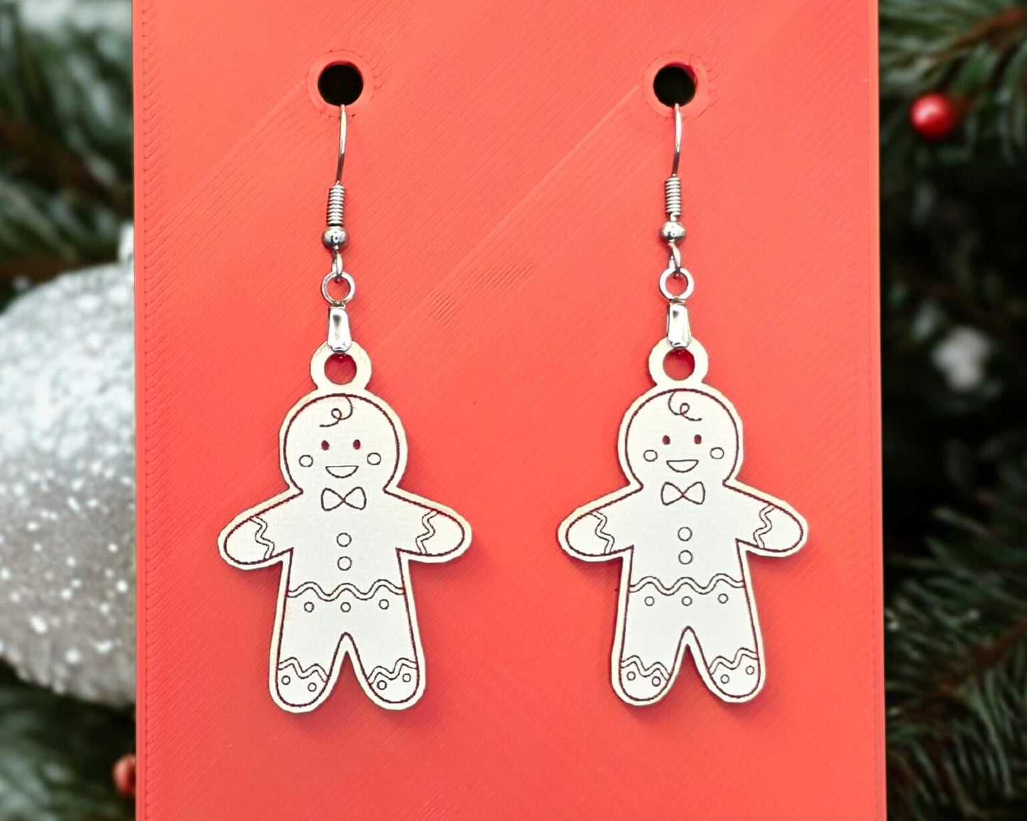 Close-up of white acrylic gingerbread man earrings hanging from nickel-free stainless-steel hooks against a bright red background. Each earring is laser-engraved with smiling gingerbread details and bow ties, with a blurred evergreen and ornament in the background. Handmade Christmas earrings by Rainbow Charlie.