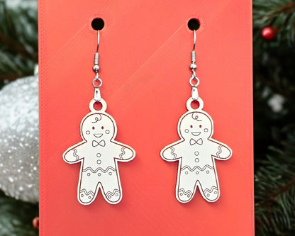 Close-up of white acrylic gingerbread man earrings hanging from nickel-free stainless-steel hooks against a bright red background. Each earring is laser-engraved with smiling gingerbread details and bow ties, with a blurred evergreen and ornament in the background. Handmade Christmas earrings by Rainbow Charlie.