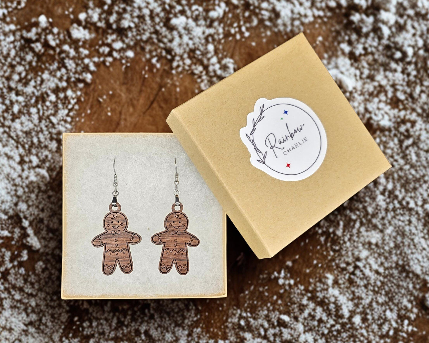 Kraft jewelry box with Rainbow Charlie logo sticker shown beside open box containing a pair of walnut gingerbread man earrings. Box rests on a wooden background sprinkled with powdered sugar for a festive, handmade presentation.
