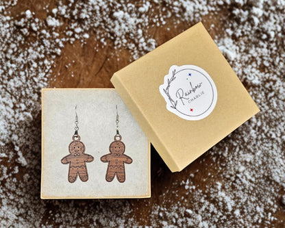 Kraft jewelry box with Rainbow Charlie logo sticker shown beside open box containing a pair of walnut gingerbread man earrings. Box rests on a wooden background sprinkled with powdered sugar for a festive, handmade presentation.