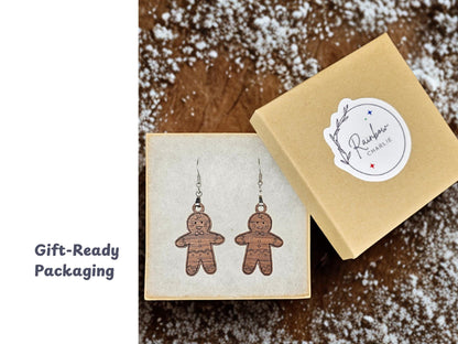 Open kraft jewelry box displaying a pair of walnut wood gingerbread man earrings on a soft white insert. A matching lid with the Rainbow Charlie logo sticker rests beside it on a dusting of powdered sugar surface. Caption reads “Gift-Ready Packaging.”