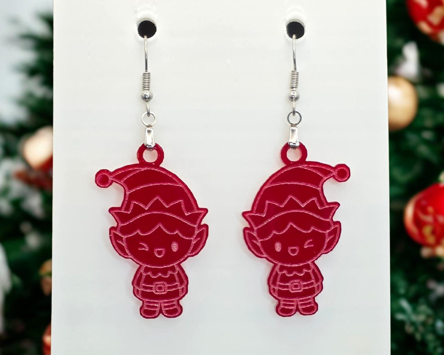 Front view of bright red acrylic elf earrings with engraved line details, hanging from nickel-free hooks on a white display card.