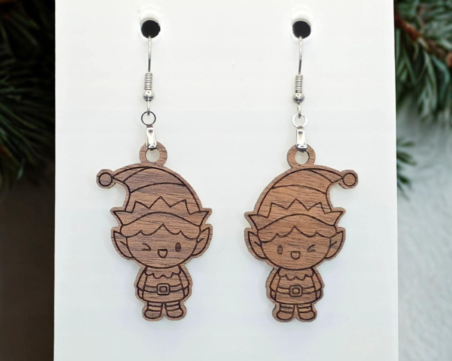 Front view of walnut wood elf earrings with laser-engraved dark details on natural grain, hanging from nickel-free stainless steel hooks on a white display card.