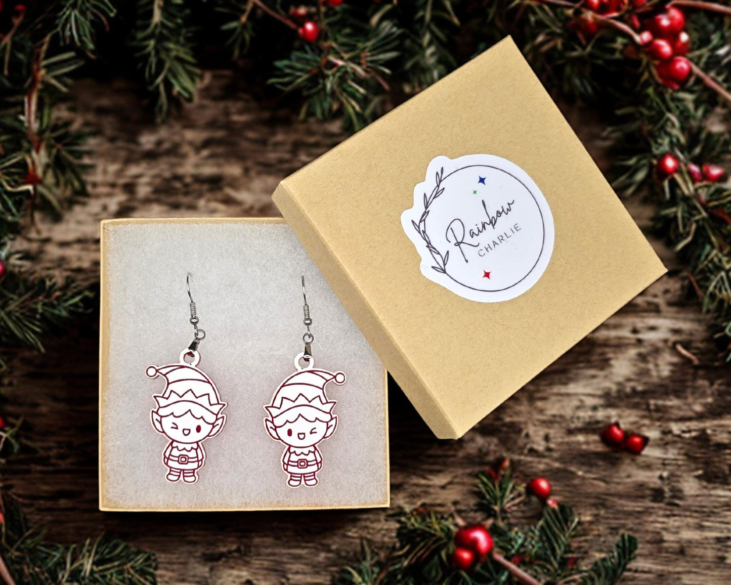 Elf earrings displayed inside a kraft jewelry gift box lined with soft white padding. The box lid, shown beside it, features a Rainbow Charlie logo sticker. Surrounding holiday greenery adds festive styling.