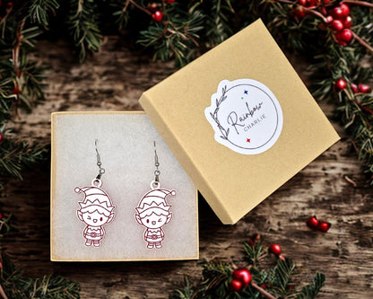 Elf earrings displayed inside a kraft jewelry gift box lined with soft white padding. The box lid, shown beside it, features a Rainbow Charlie logo sticker. Surrounding holiday greenery adds festive styling.