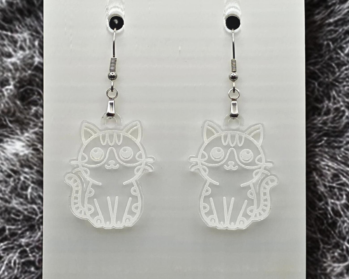 Clear acrylic cat earrings with engraved cat outline, shown on a white display card. Gray fur-like textured material appears along the left and right edges.