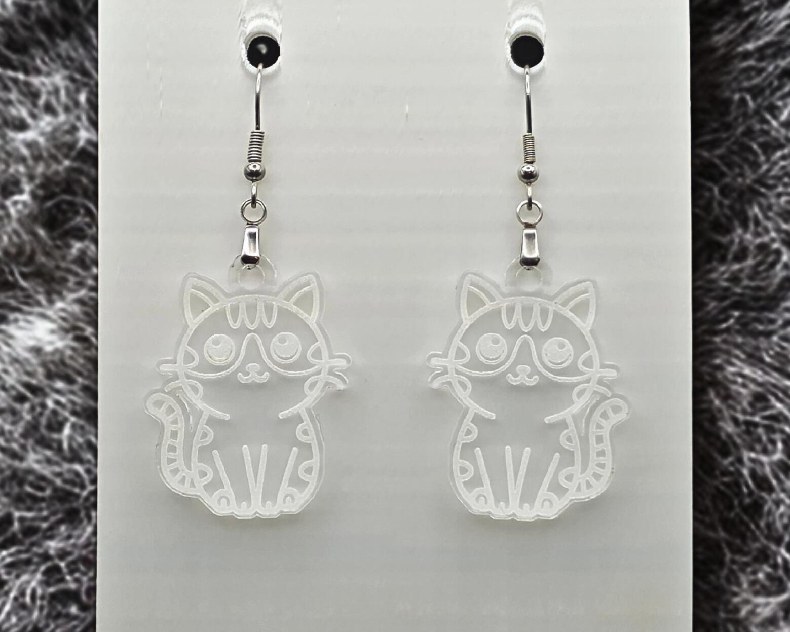 Clear acrylic cat earrings with engraved cat outline, shown on a white display card. Gray fur-like textured material appears along the left and right edges.