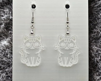 Clear acrylic cat earrings with engraved cat outline, shown on a white display card. Gray fur-like textured material appears along the left and right edges.