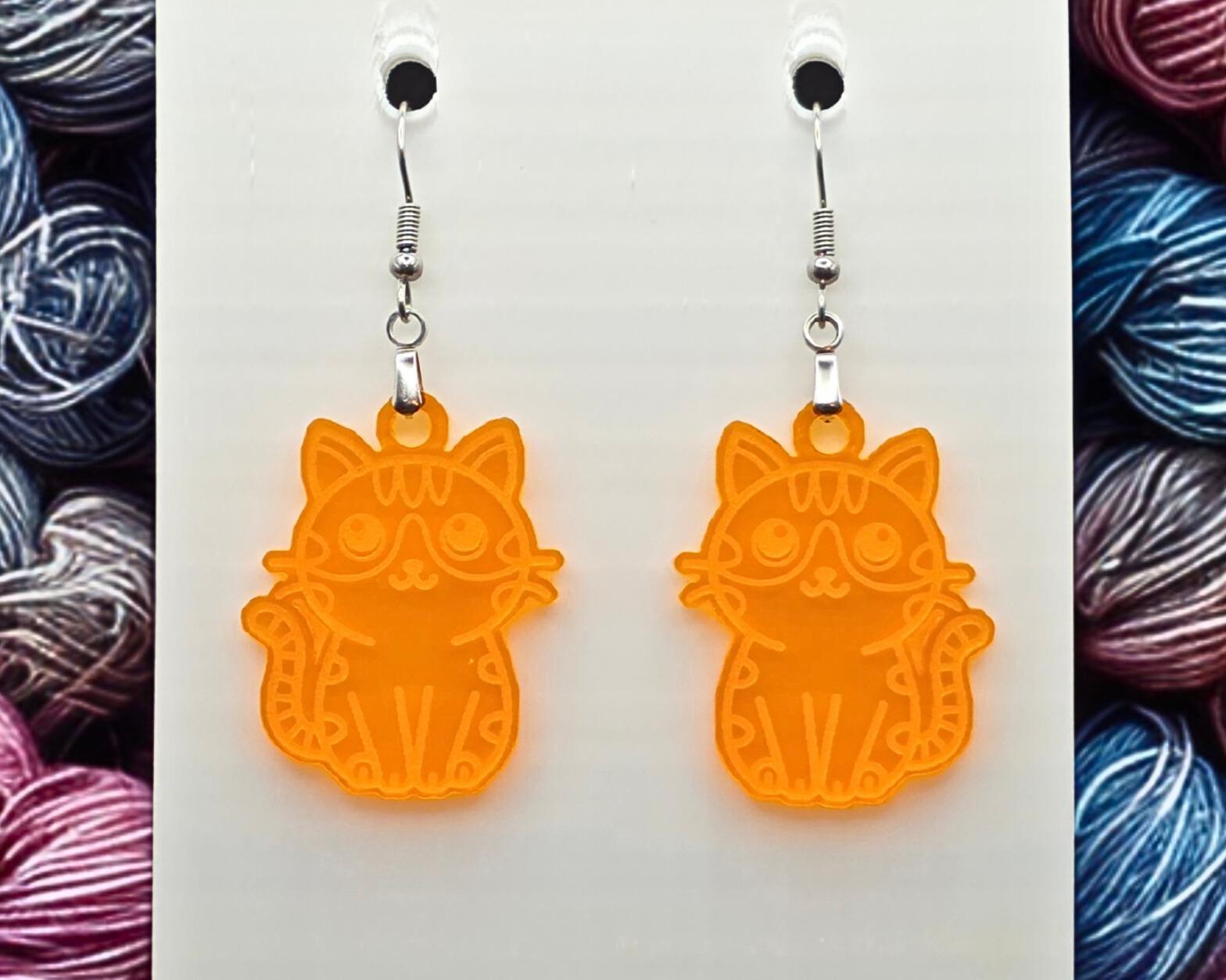 Pair of orange acrylic cat earrings with engraved details, hanging from nickel-free hooks on a white display card. Multicolored yarn balls frame the left and right sides of the image.