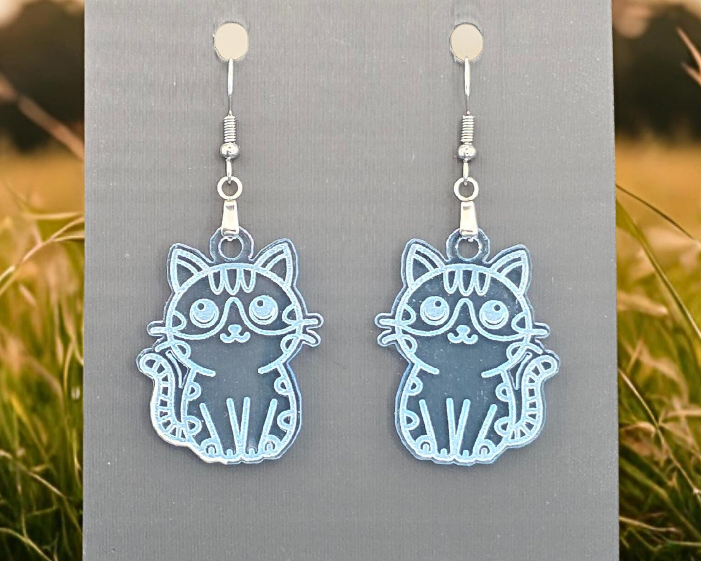 Clear blue acrylic cat earrings with engraved details displayed on a gray backing card. A field of grass appears blurred on the left and right edges.