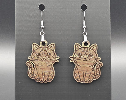 Engraved walnut wood cat earrings hanging on nickel-free hooks against a dark gray display background. The natural wood grain and engraved cat lines are clearly visible.