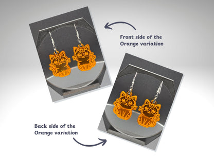 Front and back view of orange acrylic cat earrings displayed on a jewelry stand. The front side has a bright, engraved cat design, and the back shows a reversed engraving. Arrows and labels identify the front and back of the orange variation.