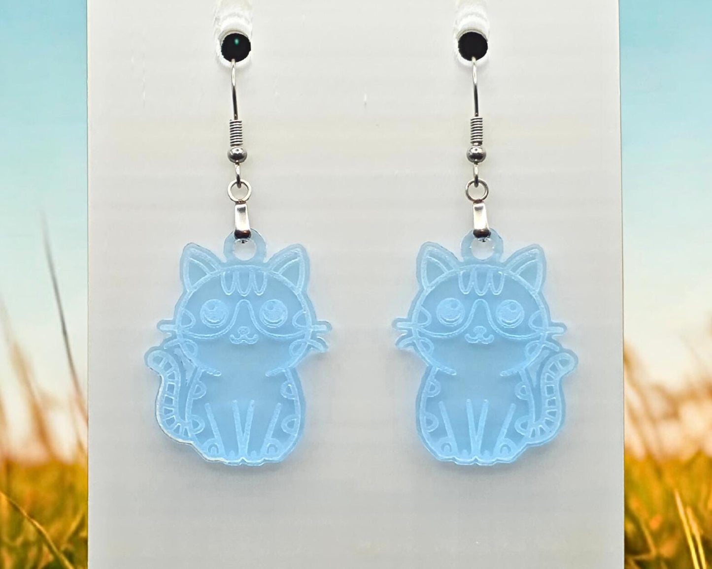 Clear blue acrylic cat earrings with engraved cat illustration, hanging on a white display card. Blurred grass and sky imagery frames the sides of the photo.