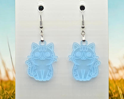 Clear blue acrylic cat earrings with engraved cat illustration, hanging on a white display card. Blurred grass and sky imagery frames the sides of the photo.