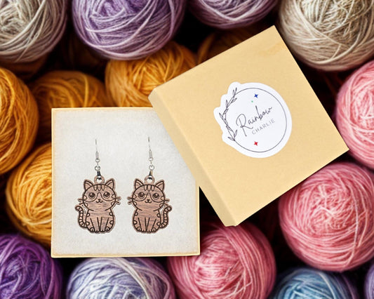 Pair of walnut wood cat earrings inside a cream-colored gift box with a Rainbow Charlie branded sticker on the lid. Colorful yarn balls fill the background.