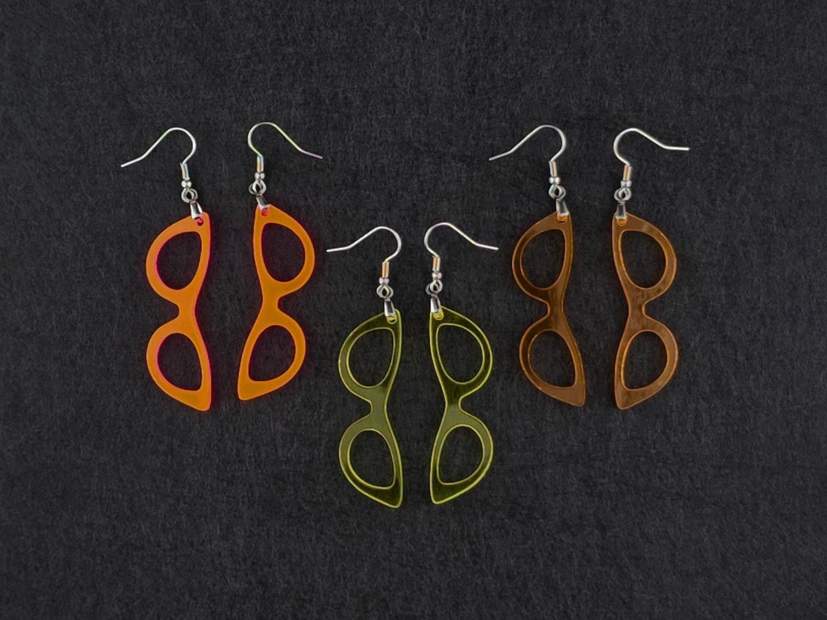Three pairs of cat-eye earrings on charcoal in fluorescent orange-pink, translucent yellow, and amber orange acrylic, showing three colorways side by side.