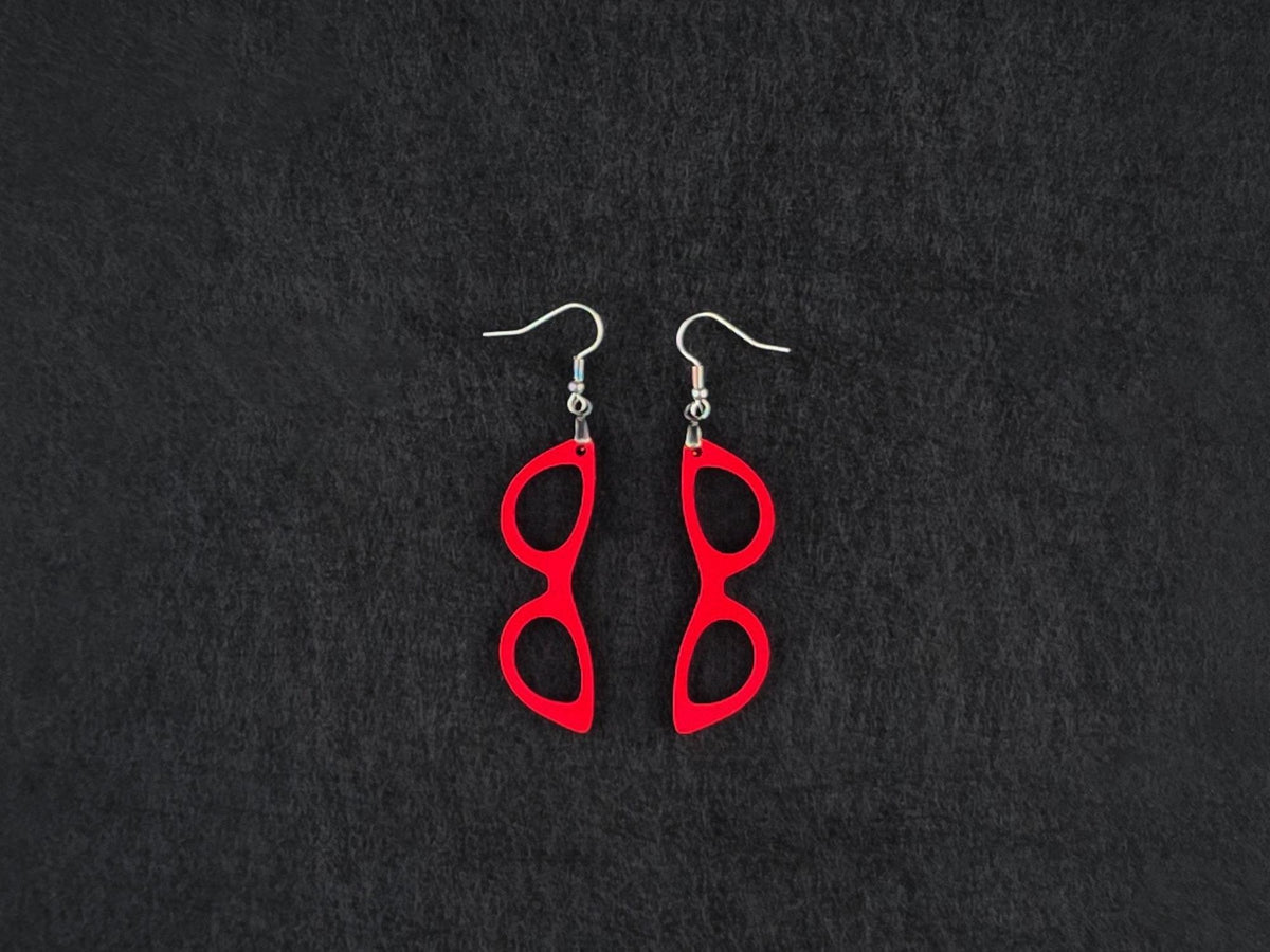 Pair of red acrylic cat-eye glasses-shaped earrings on stainless steel ear wires, flat lay on a charcoal background.