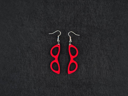 Pair of red acrylic cat-eye glasses-shaped earrings on stainless steel ear wires, flat lay on a charcoal background.