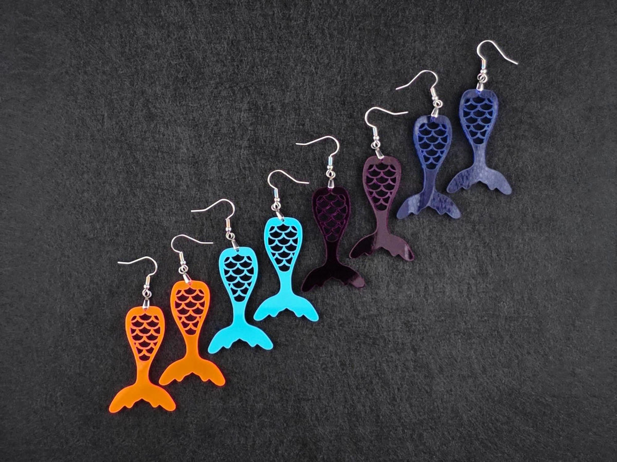 Four pairs of acrylic mermaid tail earrings in fluorescent, teal, deep purple, and blue, arranged diagonally on a charcoal background.