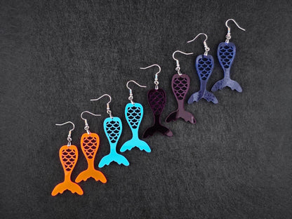 Four pairs of acrylic mermaid tail earrings in fluorescent, teal, deep purple, and blue, arranged diagonally on a charcoal background.