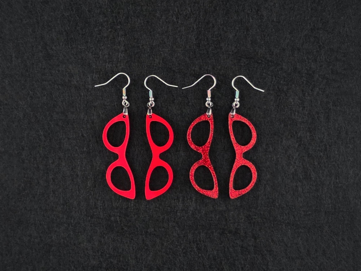 Two pairs of cat-eye earrings side by side on charcoal, one in solid red acrylic and one in a red glitter finish, showing the difference between the two textures.