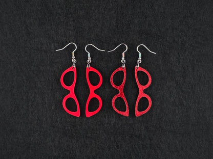Two pairs of cat-eye earrings side by side on charcoal, one in solid red acrylic and one in a red glitter finish, showing the difference between the two textures.