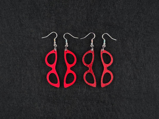 Two pairs of cat-eye earrings side by side on charcoal, one in solid red acrylic and one in a red glitter finish, showing the difference between the two textures.