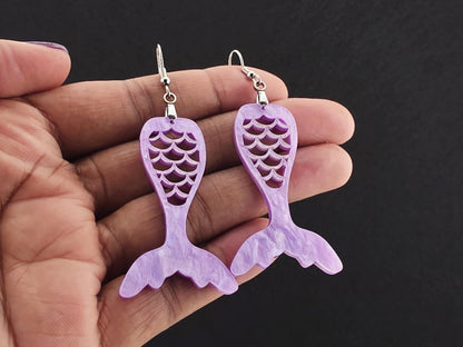 Pair of lavender pearl (marbled) acrylic mermaid tail earrings held in an open hand for scale, against a charcoal background.